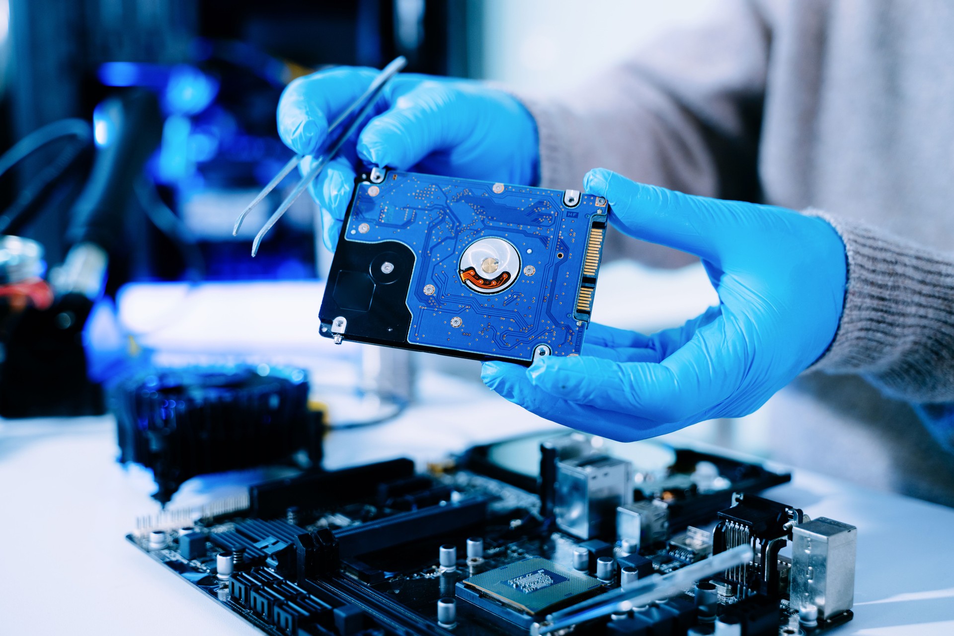 Closeup of technician with gloves holding blue hard drive circuit board above motherboard, symbolizing data recovery, storage, and computer repair.